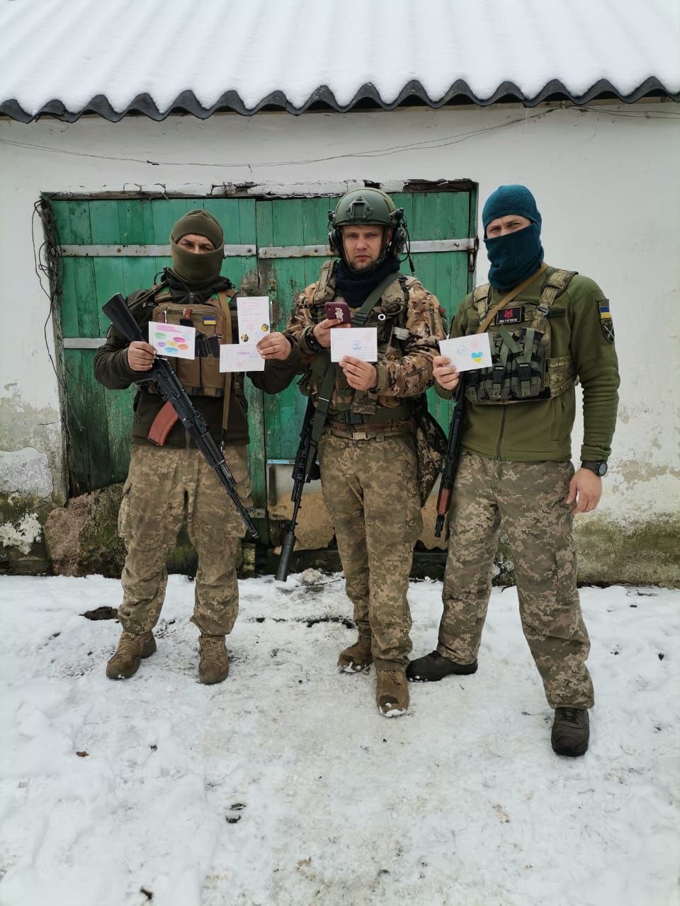 Three Ukrainian soldiers in camouflage and body armour hold up hand-decorated postcards outside a snow-covered shed