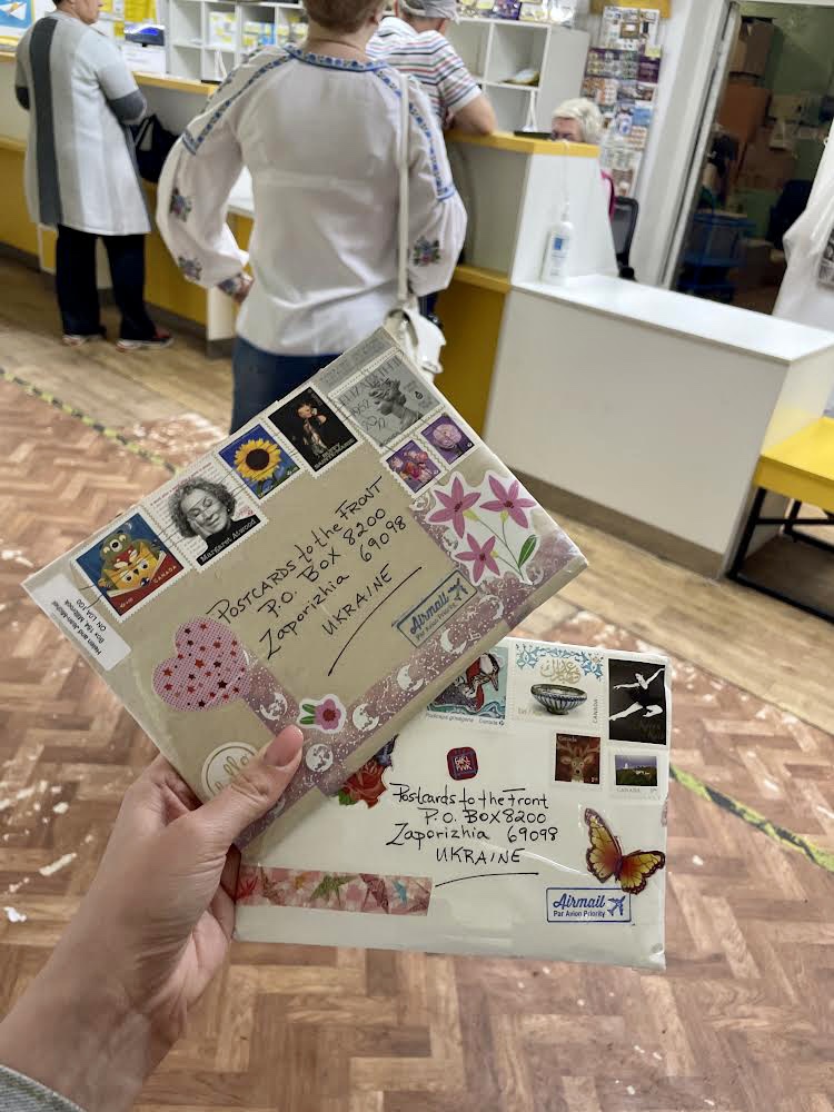 A hand holds two decorated airmail envelopes addressed to Postcards to the Front, Zaporizhzhia, Ukraine, inside a post office
