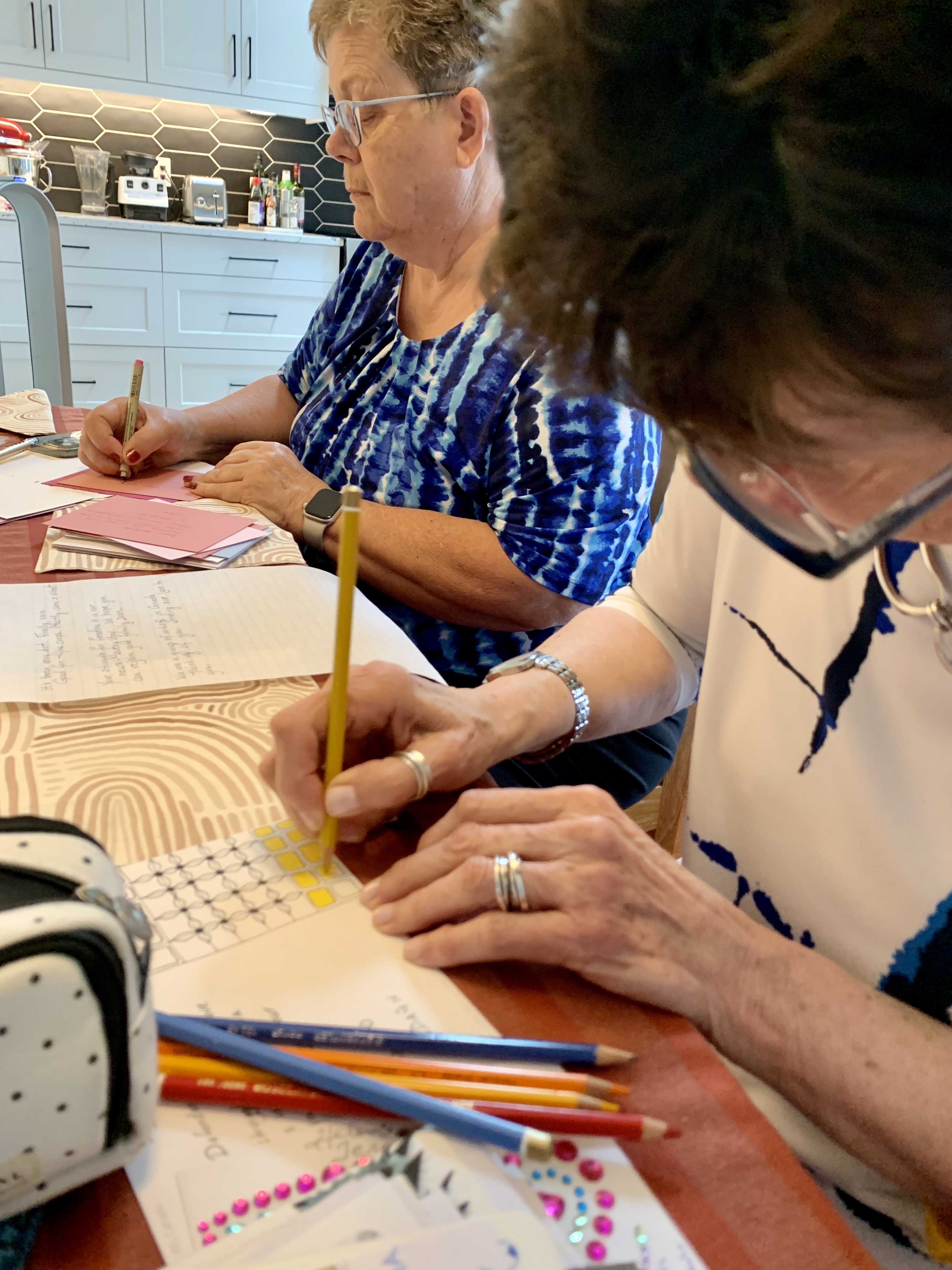Two women at a kitchen table drawing and colouring postcards with coloured pencils and craft supplies