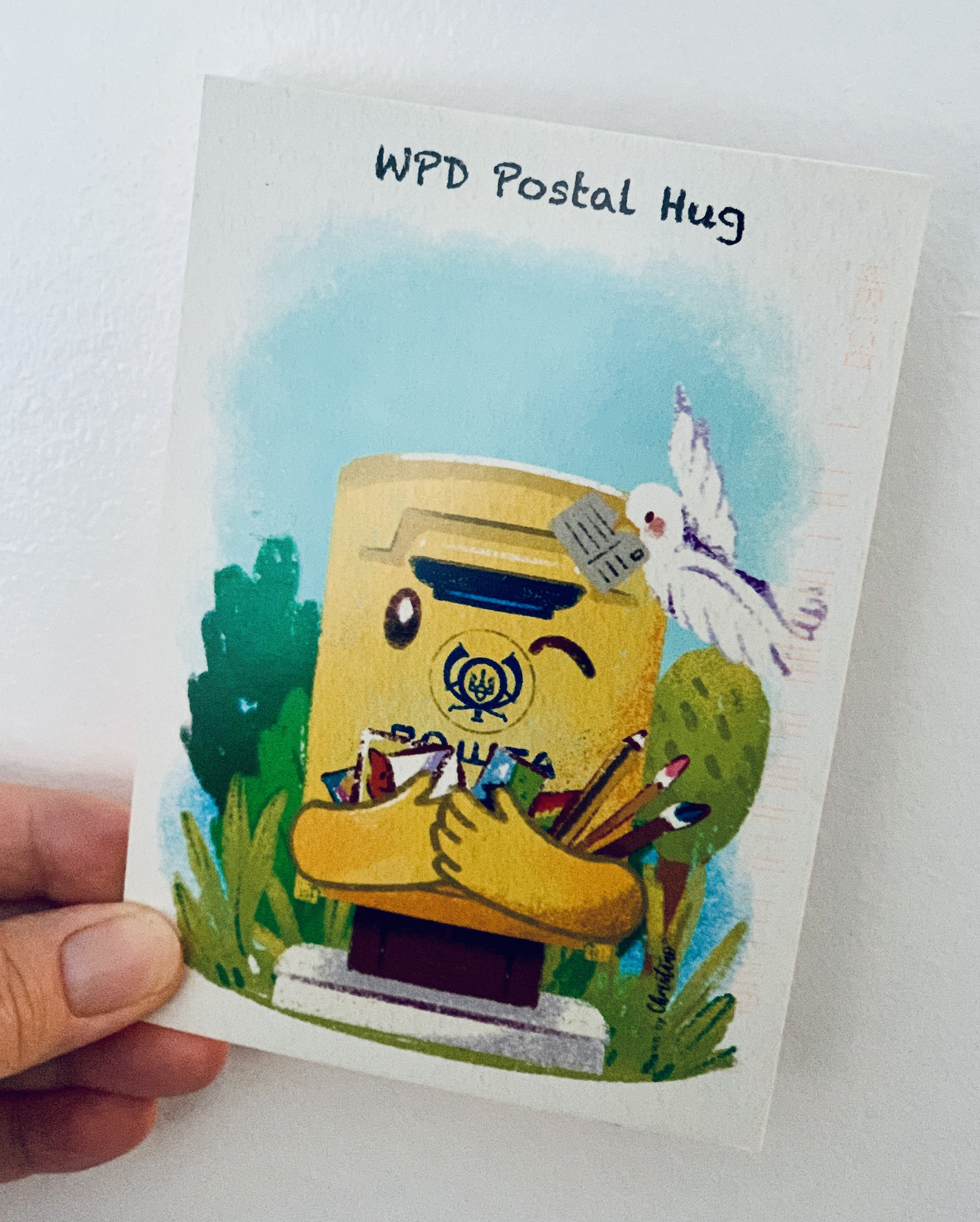 A hand holds a WPD Postal Hug postcard: a yellow Ukrainian mailbox hugging itself, with letters and a dove