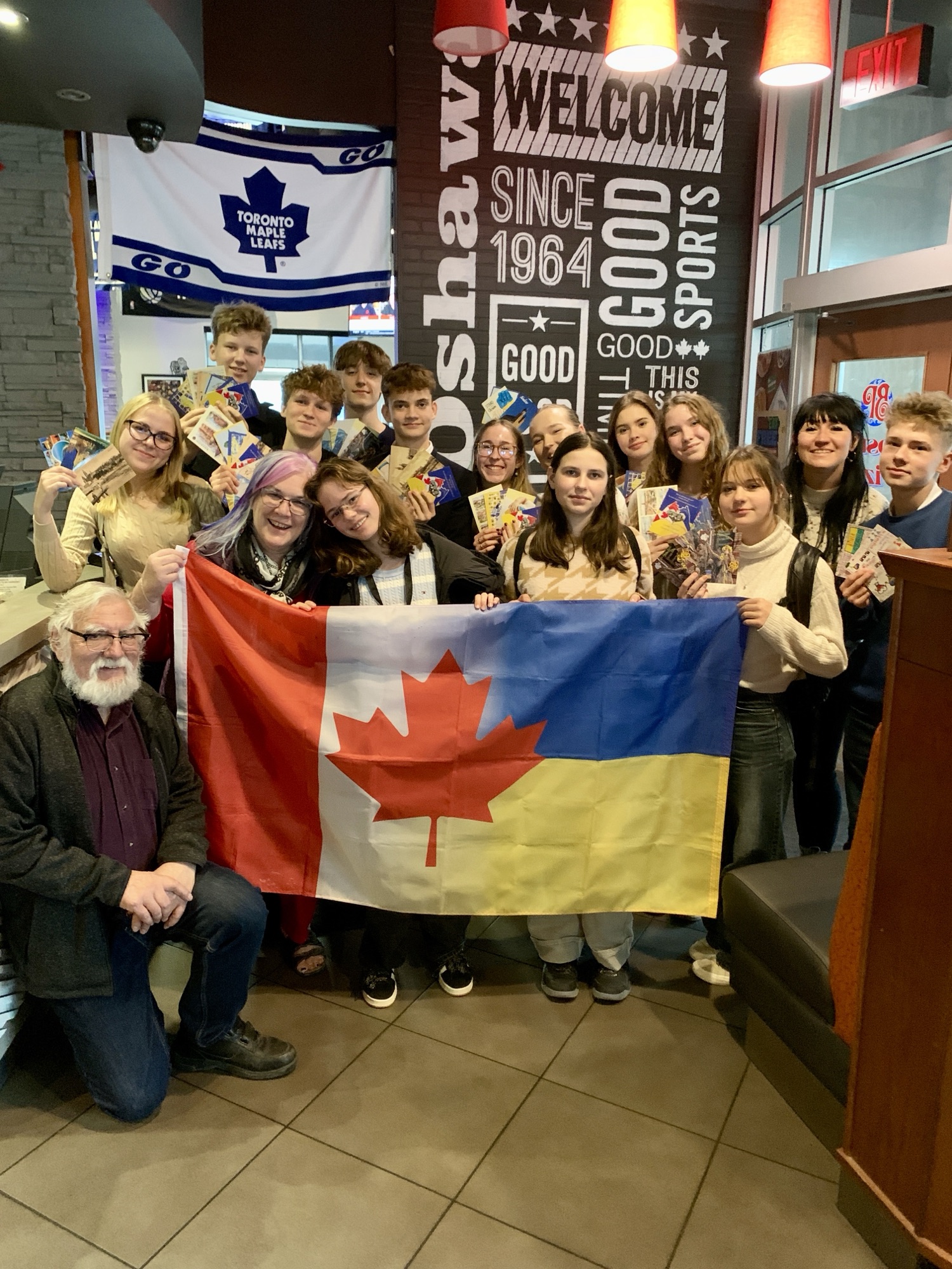 Teens and adults hold postcards behind a combined Canada-Ukraine flag inside a restaurant