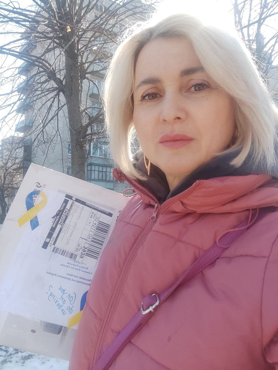 A woman in a pink coat holds a parcel tied with a blue-and-yellow ribbon on a snowy street