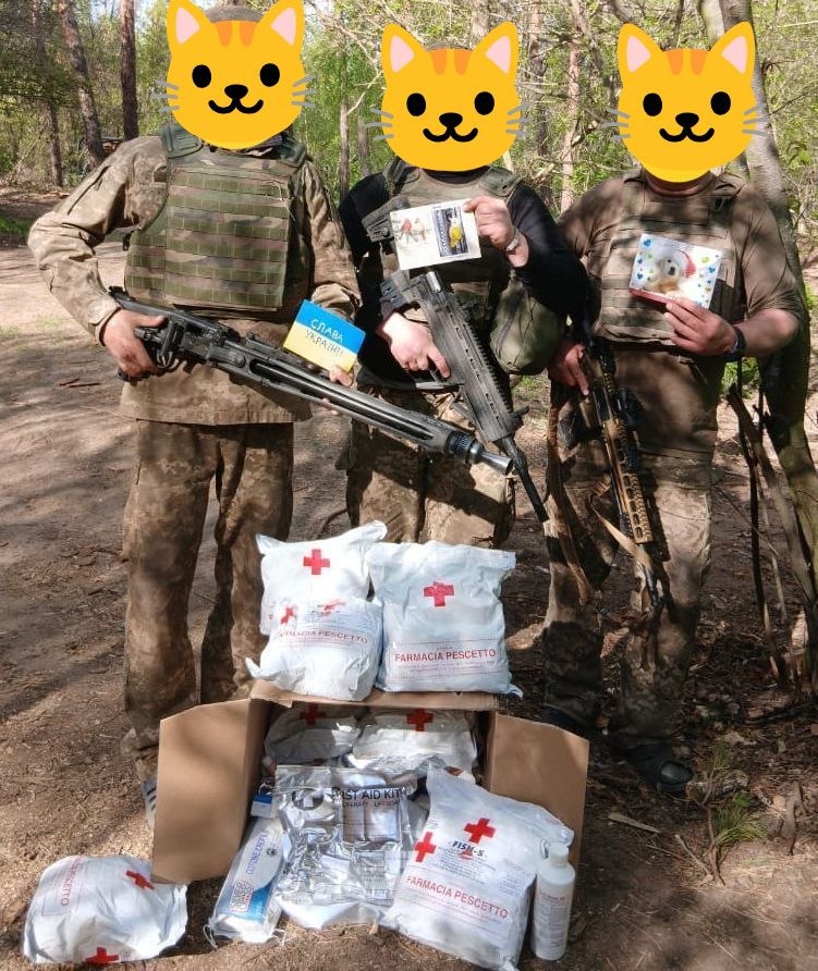 Three armed soldiers, faces hidden by cat emojis, hold postcards; one reads "Слава Україні", first-aid kits at their feet