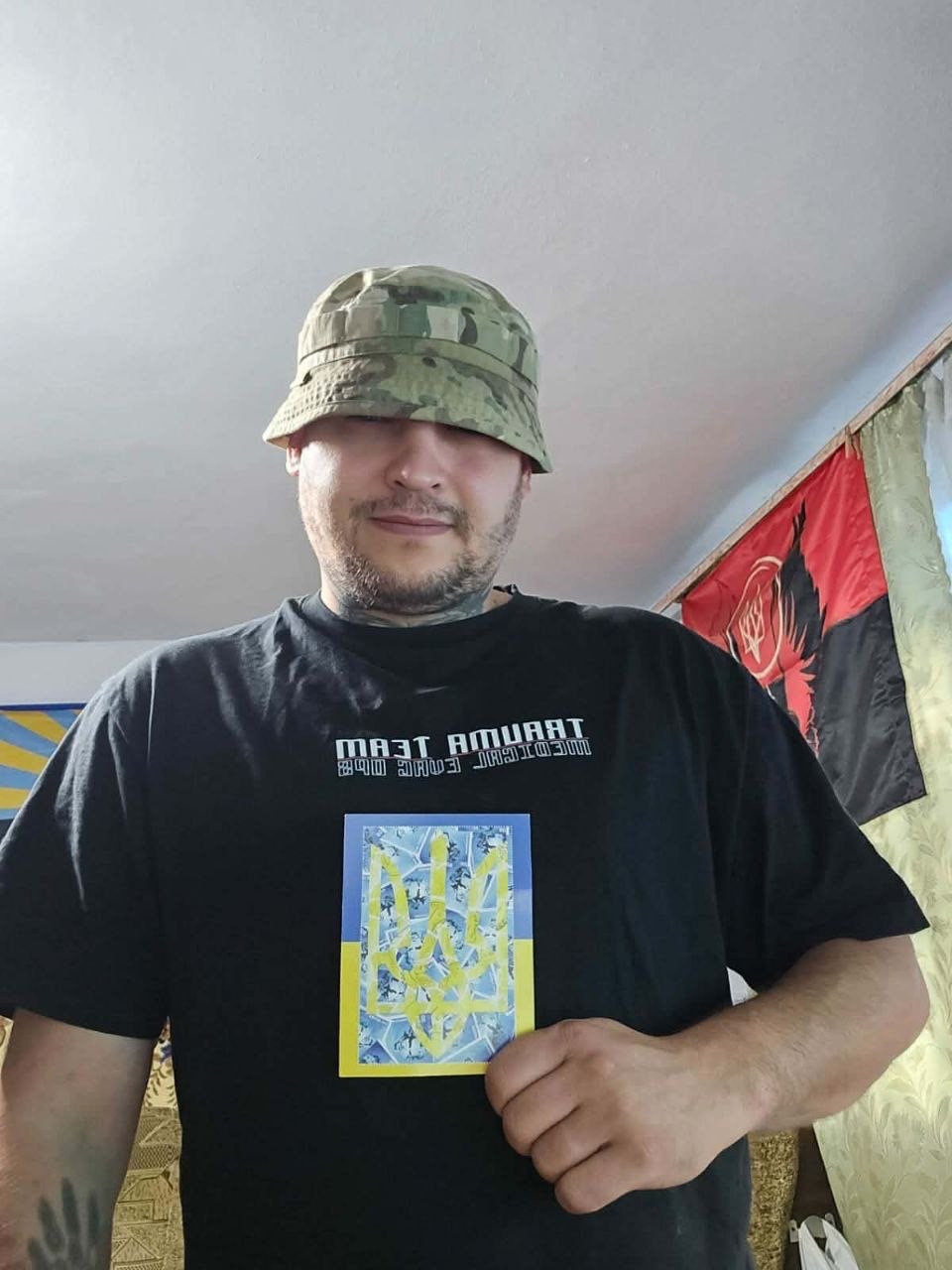 A soldier in a camouflage bucket hat holds a postcard showing a yellow tryzub over blue and yellow bands